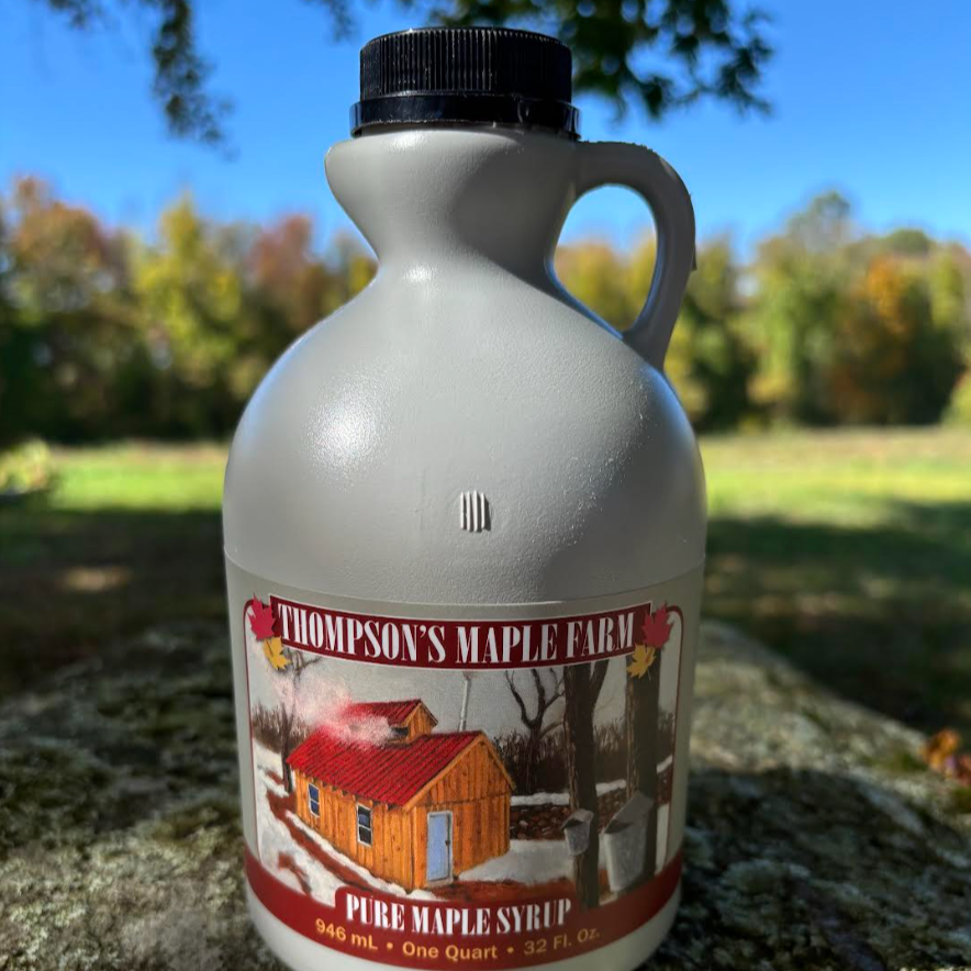 Bottle of Thompson's Maple Farm pure maple syrup, featuring a rustic cabin illustration on the label, set against a blurred outdoor background with autumn foliage. The bottle holds one quart (946 mL) of syrup, ideal for pancakes and desserts.