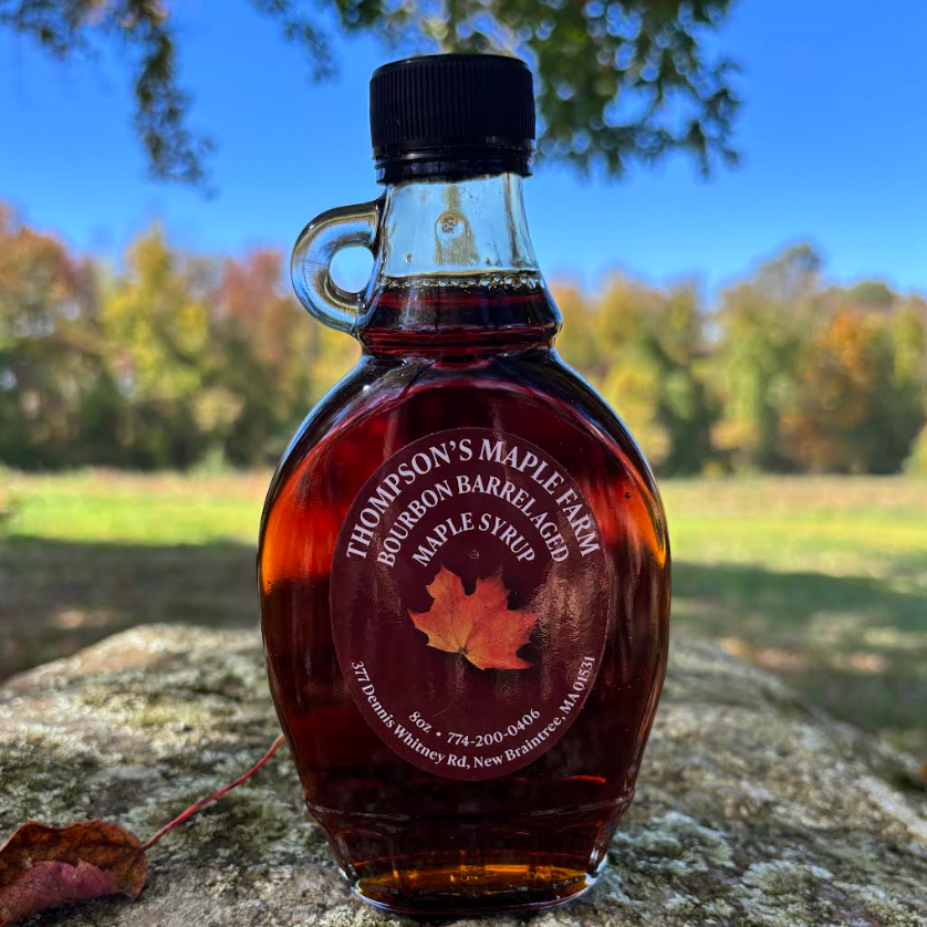 Bourbon barrel-aged maple syrup from Thompson's Maple Farm in a decorative glass bottle, set against a vibrant autumn landscape with colorful foliage in the background.