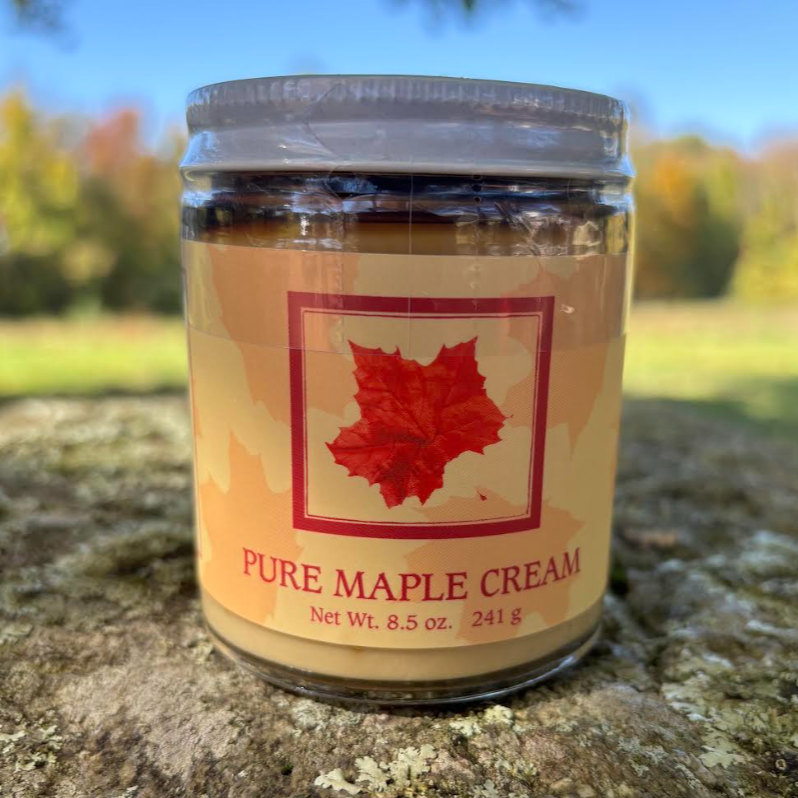 Glass jar of pure maple cream, 8.5 oz, featuring a red maple leaf design on the label, set against a blurred outdoor background with autumn foliage.